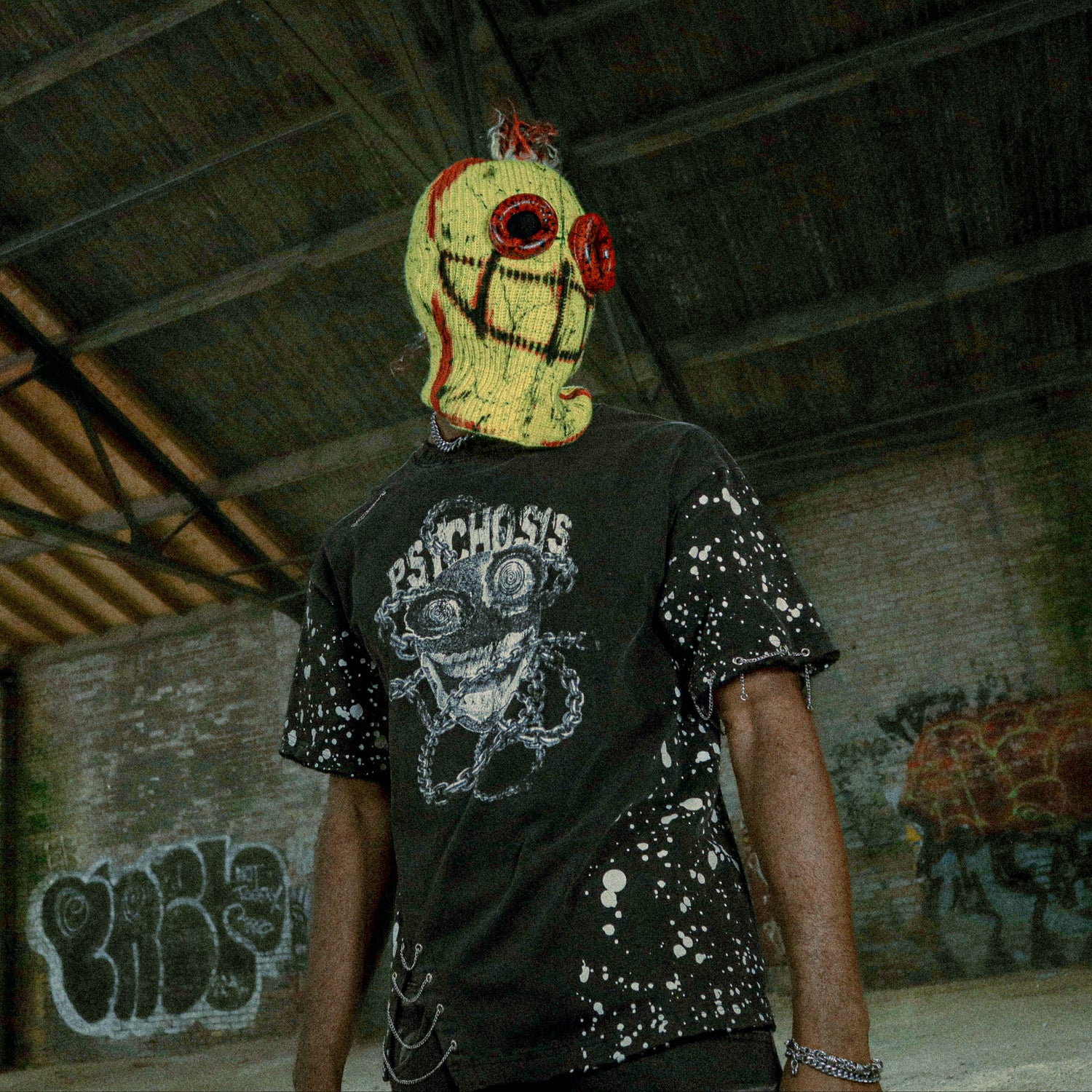 Person wearing a mask and black t-shirt with graphic design in an indoor setting with graffiti.