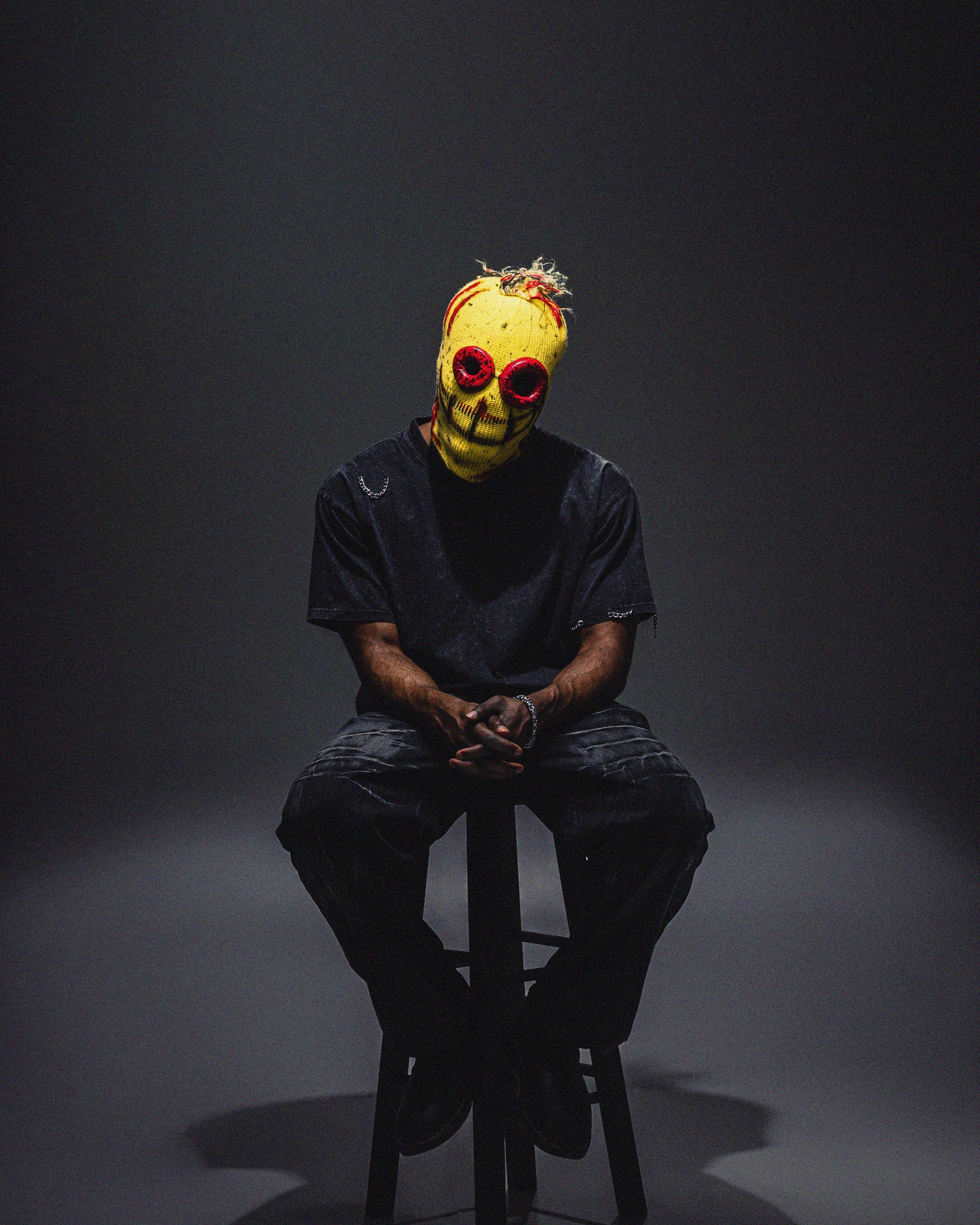 Person wearing a yellow mask with red eyes sitting on a stool against a dark background